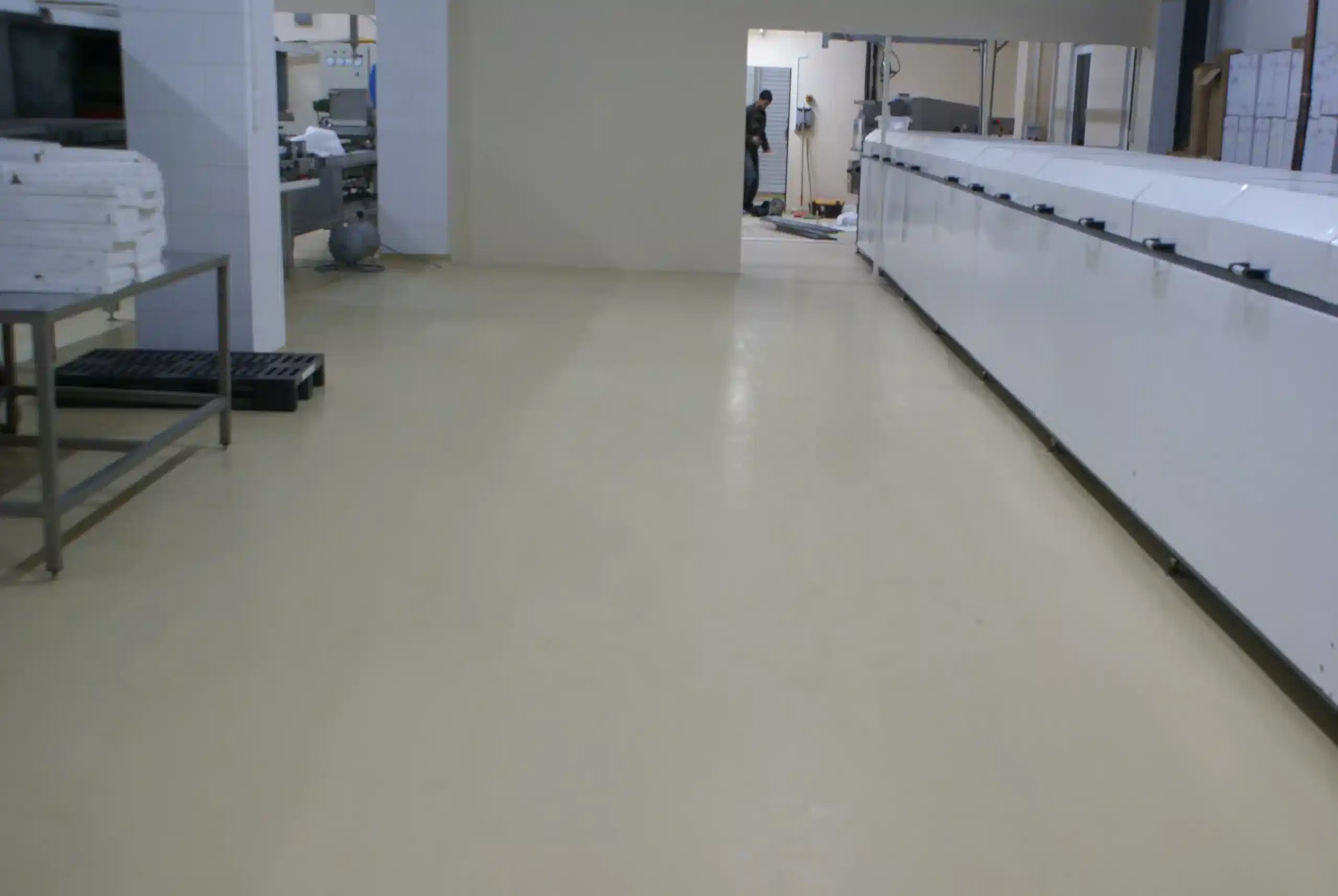 Mikrobeton Fabrika Zemini Zemini mikrobetonla yapılmış bir çikolata fabrikası. Sağda beyaz bir çikolata makinesi mevcut. Mikrobeton zeminli uzun bir koridor görünüyor.
